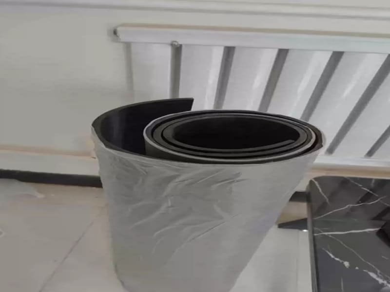 A hand using a utility knife to cut rubber flooring from the back, using a cardboard template as a guide.